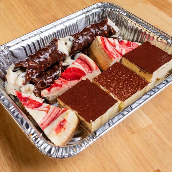 A foil tray filled with cannolis, tiramisu, and cheesecake slices on a wooden table.