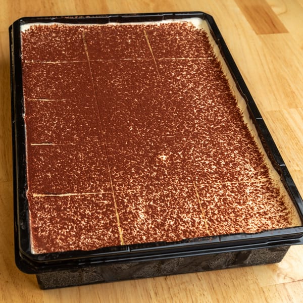 A large tray of tiramisu, dusted with cocoa powder and pre-cut into squares, on a wooden table.