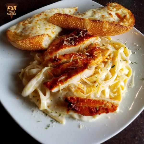 Creamy fettuccine alfredo with sliced chicken and two pieces of cheesy garlic bread on a white plate.
