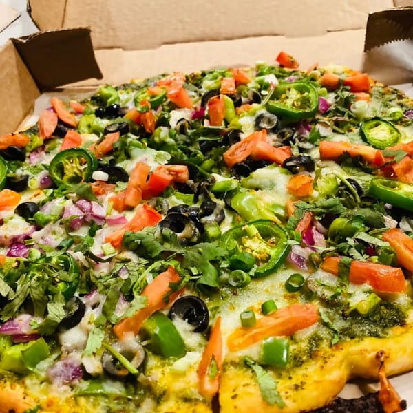 A Fun Pizza Kitchen pizza with green sauce, tomatoes, olives, jalapenos, and onions in a box.
